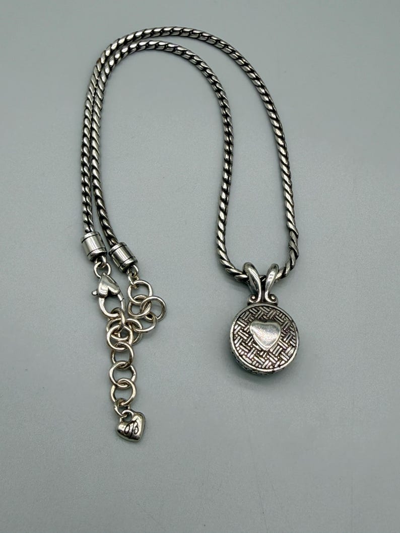 May include: A silver-toned necklace featuring a round pendant with a textured, woven-style pattern and a smooth, raised heart in the center. The chain is a thick, twisted rope style with a lobster claw clasp and an adjustable link extender.