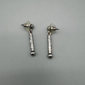 May include: A pair of silver-colored dangle earrings. Each earring features a post back and a cylindrical design with etched patterns. The bottom of each earring has a small, round, silver-colored bead.