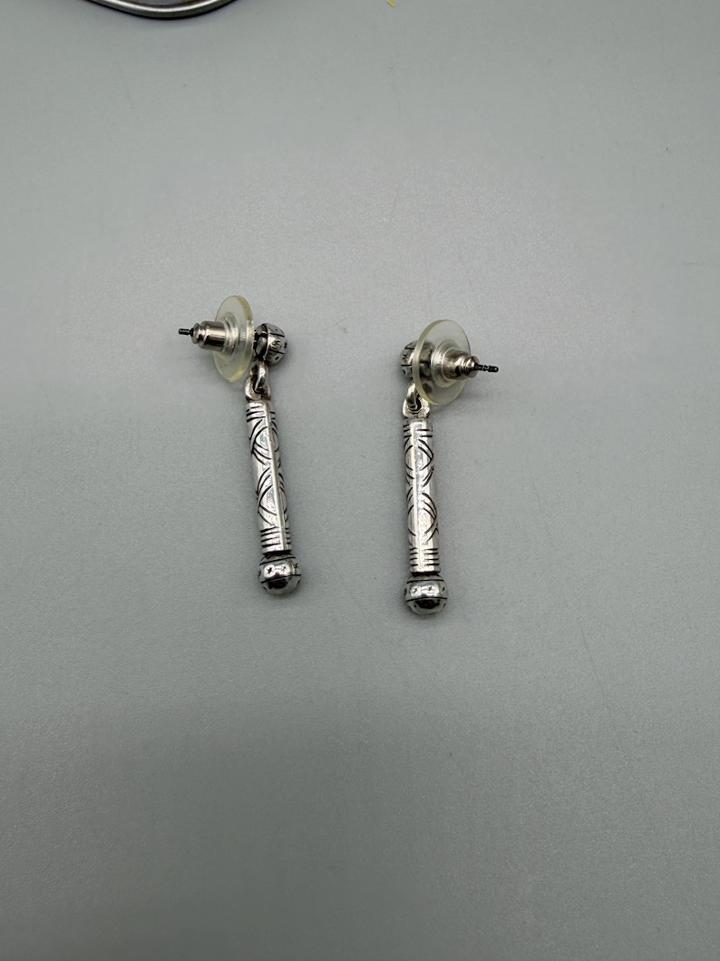May include: A pair of silver-colored dangle earrings. Each earring features a post back and a cylindrical design with etched patterns. The bottom of each earring has a small, round, silver-colored bead.