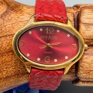 May include: A Joan Rivers Classics quartz watch with an oval gold-tone case and a deep red sunray dial. The dial features gold-tone numerals at 12, 3, 6, and 9, with small crystal markers at other positions. It has a red woven leather strap.