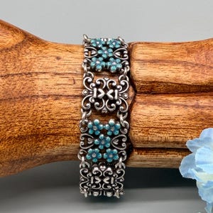以下が含まれることがあります： シルバーカラーのブレスレット。精巧なスクロールワークとターコイズブルーの花のアクセントが特徴です。ブレスレットは、小さなチェーンで接続された複数の長方形のセグメントで構成されています。各セグメントには、小さなターコイズブルーの花のフローラルデザインがあります。