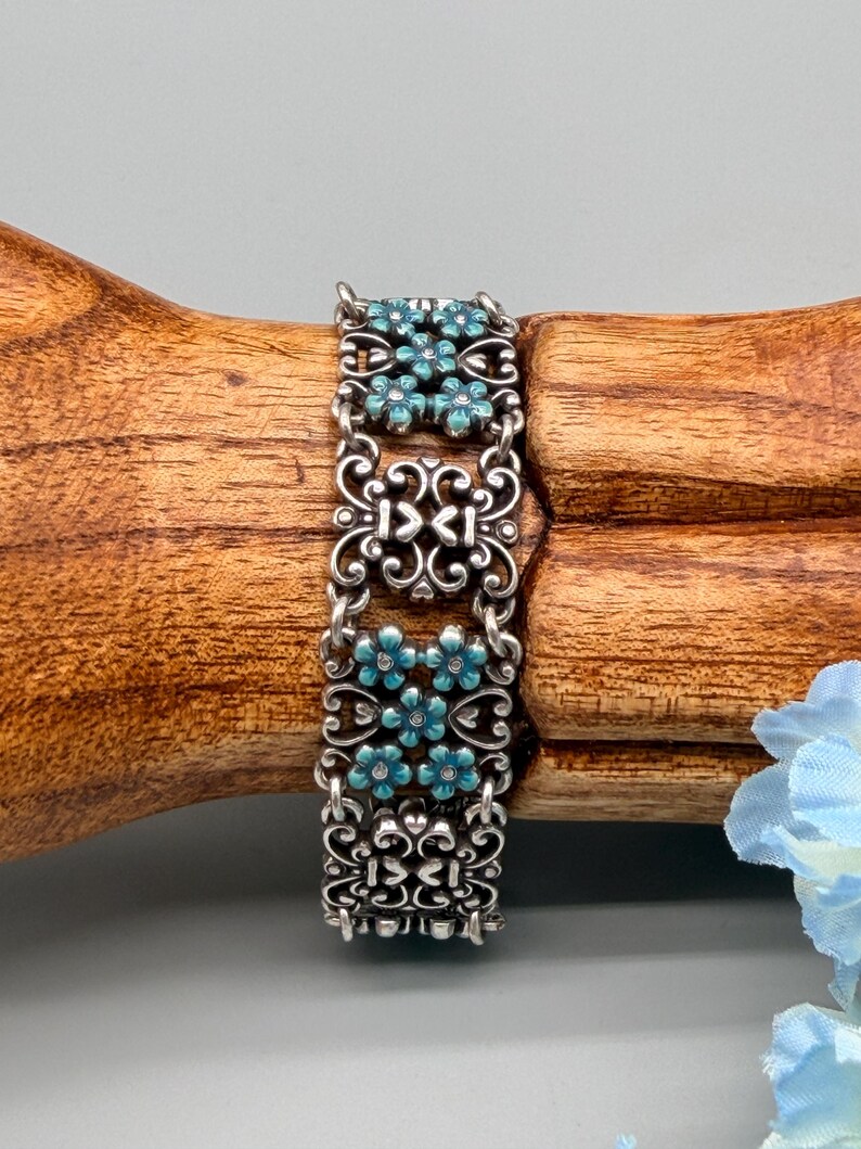 以下が含まれることがあります： シルバーカラーのブレスレット。精巧なスクロールワークとターコイズブルーの花のアクセントが特徴です。ブレスレットは、小さなチェーンで接続された複数の長方形のセグメントで構成されています。各セグメントには、小さなターコイズブルーの花のフローラルデザインがあります。