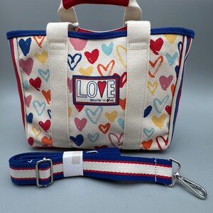 Puede incluir: Bolso tote de lona con un estampado de corazones coloridos y la palabra "LOVE" en la parte delantera. El bolso tiene un ribete azul, detalles en rojo y una correa para el hombro desmontable. Las asas son blancas con la parte superior roja.