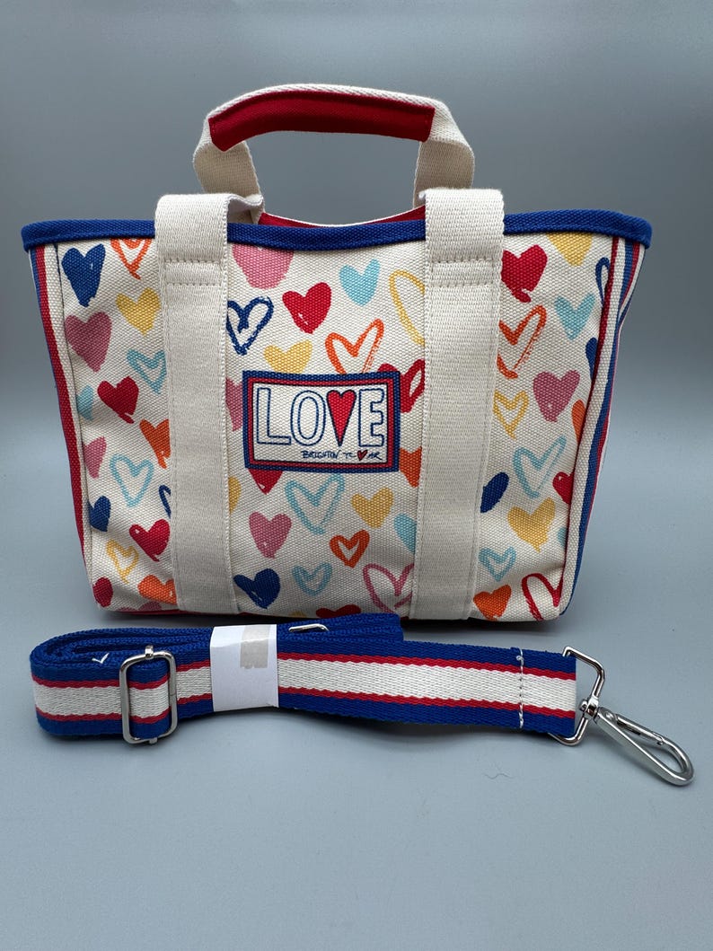 Puede incluir: Bolso tote de lona con un estampado de corazones coloridos y la palabra "LOVE" en la parte delantera. El bolso tiene un ribete azul, detalles en rojo y una correa para el hombro desmontable. Las asas son blancas con la parte superior roja.