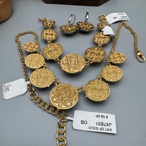 May include: A gold-toned jewelry set featuring a necklace, bracelet, and earrings. The necklace and bracelet have circular, patterned medallions. The earrings are small, round, and patterned. The set is displayed on a gray surface. The text "ART OF GOLD" is visible.