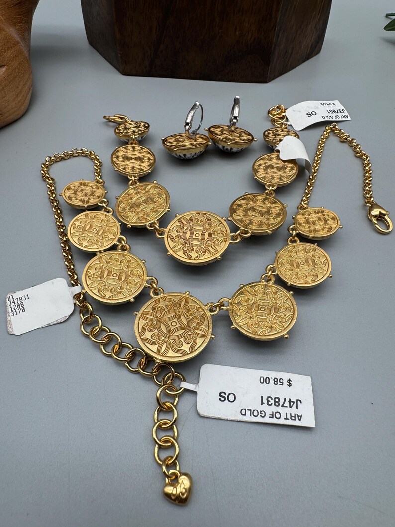 May include: A gold-toned jewelry set featuring a necklace, bracelet, and earrings. The necklace and bracelet have circular, patterned medallions. The earrings are small, round, and patterned. The set is displayed on a gray surface. The text "ART OF GOLD" is visible.