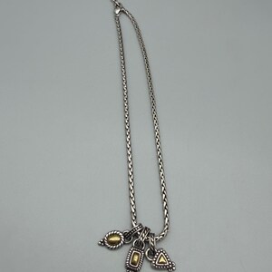 May include: A silver-toned chain necklace featuring a braided link design and an adjustable clasp. Three small charms hang from the bottom, each displaying a combination of silver-toned metal and a central gold-toned accent in oval, rectangular, and triangular shapes.