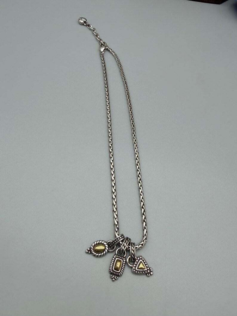 May include: A silver-toned chain necklace featuring a braided link design and an adjustable clasp. Three small charms hang from the bottom, each displaying a combination of silver-toned metal and a central gold-toned accent in oval, rectangular, and triangular shapes.