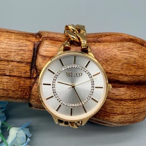 May include: A gold-toned SO & CO New York watch with a silver face and crystal accents. The watch features a chain-link band and is displayed on a wooden surface. The watch has a round face with gold hands and markers.
