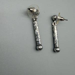May include: A pair of silver-toned dangle earrings. Each earring features a cylindrical design with etched patterns and a rounded bottom. The earrings have post-style backs with clear plastic earring stoppers.