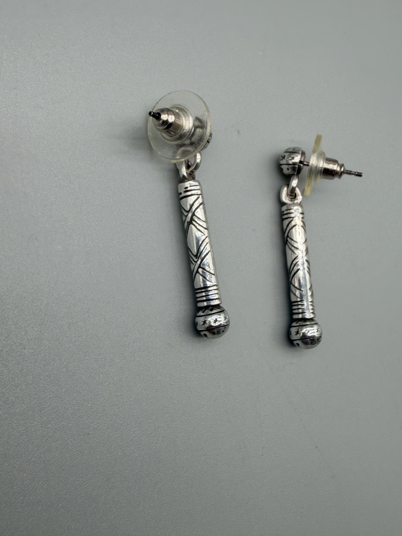 May include: A pair of silver-toned dangle earrings. Each earring features a cylindrical design with etched patterns and a rounded bottom. The earrings have post-style backs with clear plastic earring stoppers.