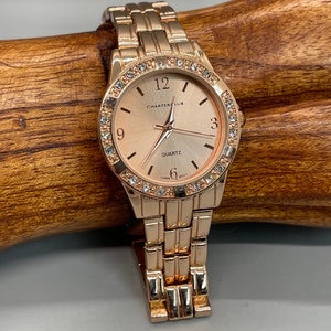 May include: A rose gold tone watch with a rose gold tone metal band. The watch face is round and has a white background with black Roman numeral hour markers. The watch face also has a small seconds dial at the 6 o'clock position. The watch face is surrounded by a row of clear crystals. The watch face has the text "Charter Club" and "Quartz" printed on it.