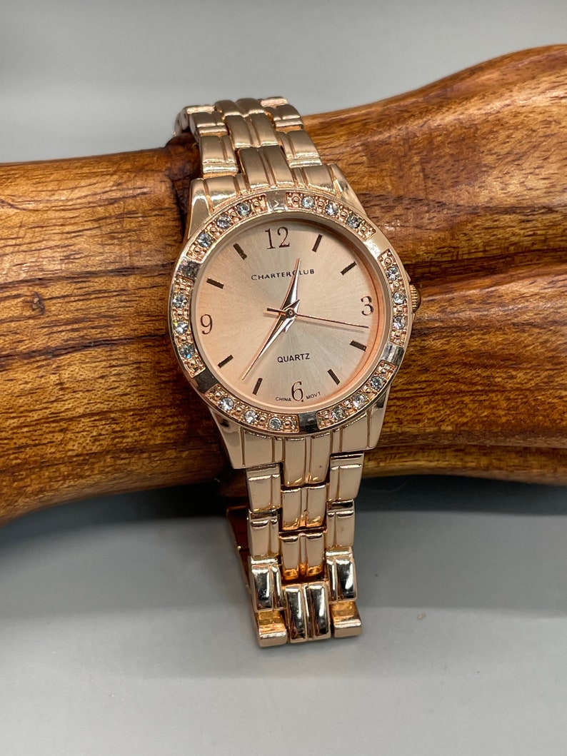 May include: A rose gold tone watch with a rose gold tone metal band. The watch face is round and has a white background with black Roman numeral hour markers. The watch face also has a small seconds dial at the 6 o'clock position. The watch face is surrounded by a row of clear crystals. The watch face has the text "Charter Club" and "Quartz" printed on it.