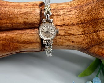 May include: A silver-toned wristwatch with a round face, featuring a light-colored dial with numerical markers at the twelve and six o'clock positions. The watch band is composed of intricate, decorative silver links set with small clear stones.