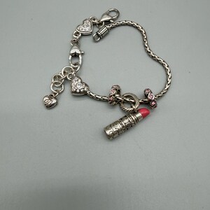 May include: A silver-toned charm bracelet featuring a braided chain, a lipstick charm with a pink tip, two heart-shaped charms encrusted with clear crystals, two small pink-accented spacer beads, and a small dangling heart charm with an engraved design.