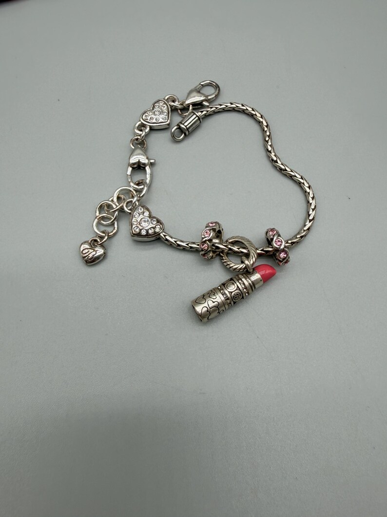 May include: A silver-toned charm bracelet featuring a braided chain, a lipstick charm with a pink tip, two heart-shaped charms encrusted with clear crystals, two small pink-accented spacer beads, and a small dangling heart charm with an engraved design.