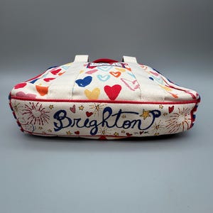 Puede incluir: Un bolso de mano color crema con ribete y asas rojas. El bolso est&aacute; decorado con corazones coloridos, fuegos artificiales y la palabra "Brighton" en cursiva. El bolso tiene forma rectangular.