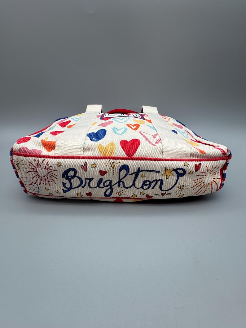 Puede incluir: Un bolso de mano color crema con ribete y asas rojas. El bolso est&aacute; decorado con corazones coloridos, fuegos artificiales y la palabra "Brighton" en cursiva. El bolso tiene forma rectangular.