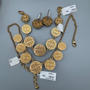May include: A gold-toned jewelry set featuring a necklace, earrings, and bracelet. The necklace and bracelet are composed of circular, patterned medallions. The earrings have a similar design. The set is displayed on a gray surface.