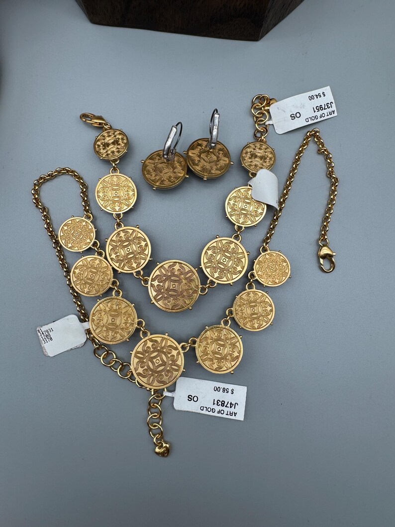 May include: A gold-toned jewelry set featuring a necklace, earrings, and bracelet. The necklace and bracelet are composed of circular, patterned medallions. The earrings have a similar design. The set is displayed on a gray surface.