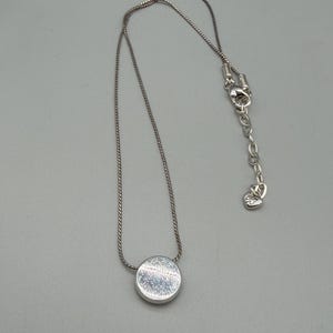 Puede incluir: Un collar de plata con un colgante circular. El colgante tiene una superficie texturizada y est&aacute; unido a una delicada cadena. El collar tiene un cierre ajustable. La joya se muestra sobre un fondo gris.