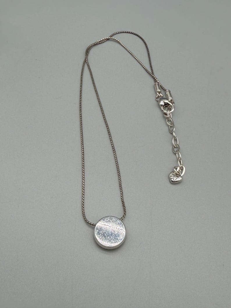 Puede incluir: Un collar de plata con un colgante circular. El colgante tiene una superficie texturizada y est&aacute; unido a una delicada cadena. El collar tiene un cierre ajustable. La joya se muestra sobre un fondo gris.