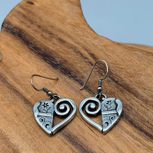 May include: A pair of silver-toned heart-shaped earrings. Each earring features a floral design on one side and a swirl design on the other. The earrings have a hook closure.
