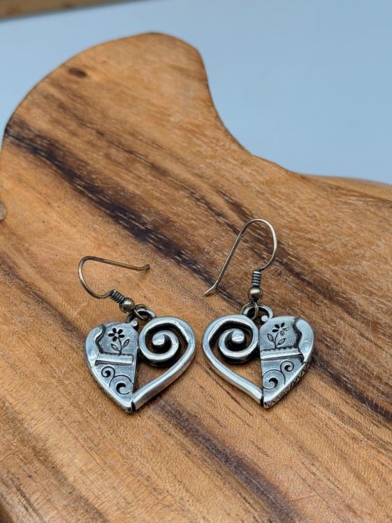 May include: A pair of silver-toned heart-shaped earrings. Each earring features a floral design on one side and a swirl design on the other. The earrings have a hook closure.