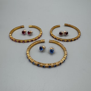 Op de afbeelding: Drie sieradensets: goudkleurige armbanden en bijpassende oorstekers. Twee sets hebben rode edelstenen, terwijl de derde set blauwe edelstenen heeft. De armbanden hebben een vierkant ontwerp.