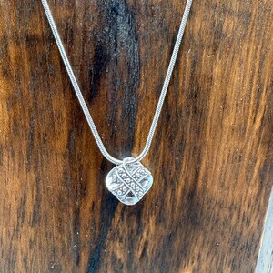 May include: A silver-toned necklace with a square pendant. The pendant has a detailed, textured design and is suspended from a thin, snake chain. The necklace is displayed against a dark wood background.