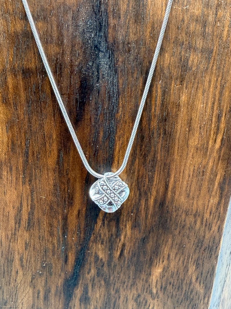 May include: A silver-toned necklace with a square pendant. The pendant has a detailed, textured design and is suspended from a thin, snake chain. The necklace is displayed against a dark wood background.