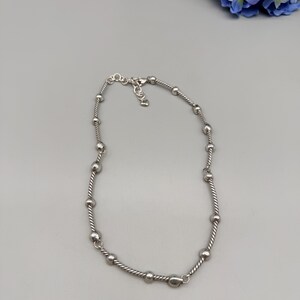 May include: A silver-toned necklace featuring a twisted rope chain design interspersed with smooth, round metal beads. The necklace includes a small metal clasp closure at the back, providing a secure and adjustable fit for the wearer.
