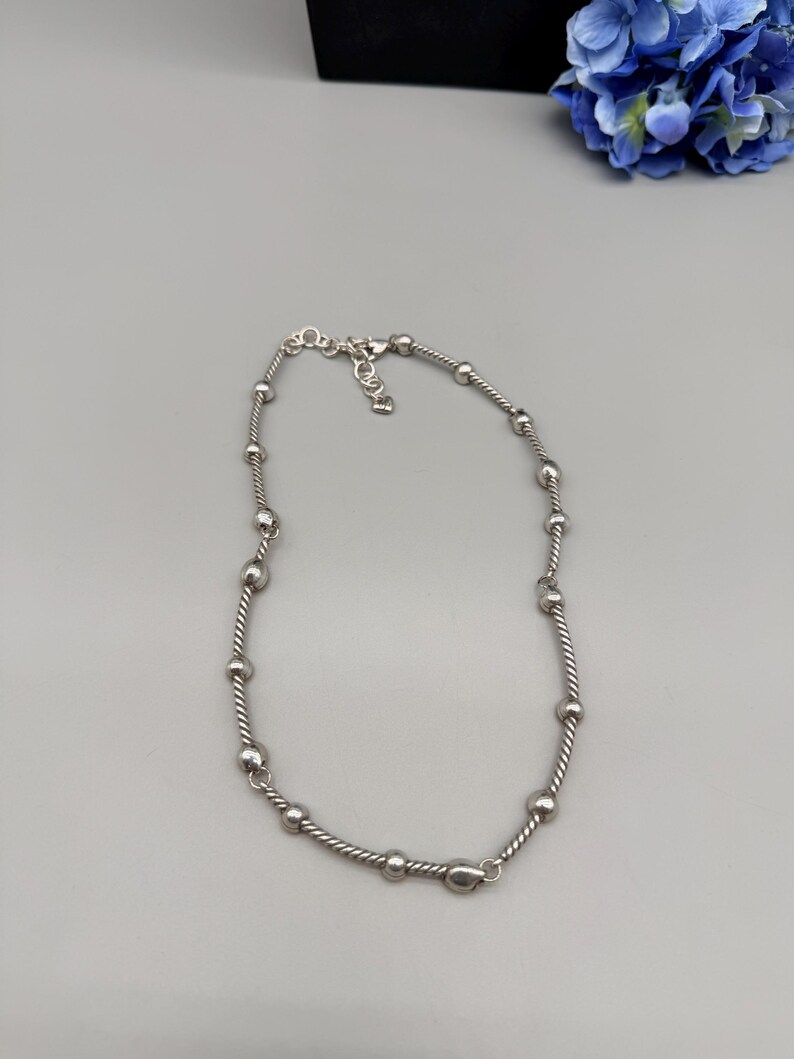 May include: A silver-toned necklace featuring a twisted rope chain design interspersed with smooth, round metal beads. The necklace includes a small metal clasp closure at the back, providing a secure and adjustable fit for the wearer.