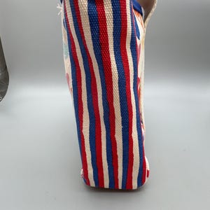 Puede incluir: Un bolso de mano de lona con rayas verticales rojas, blancas y azules. El borde superior est&aacute; ribeteado con tela azul. El bolso tiene un asa y una base rectangular. El bolso est&aacute; hecho de un material resistente.