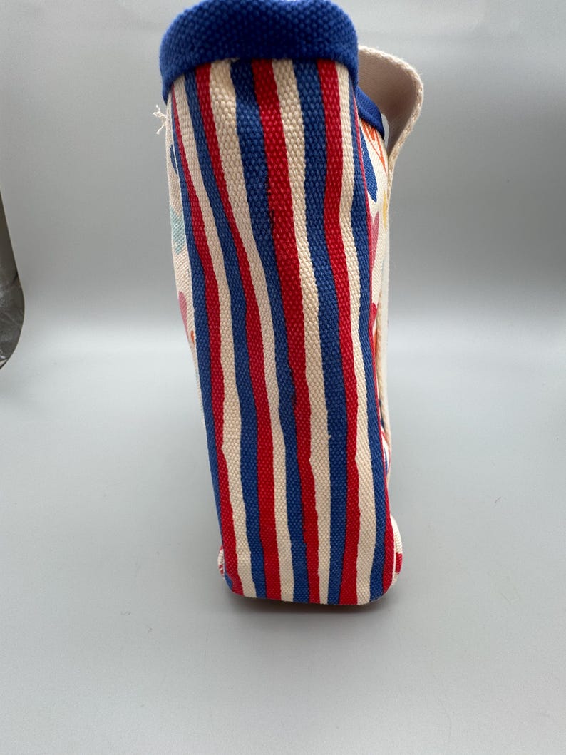 Puede incluir: Un bolso de mano de lona con rayas verticales rojas, blancas y azules. El borde superior est&aacute; ribeteado con tela azul. El bolso tiene un asa y una base rectangular. El bolso est&aacute; hecho de un material resistente.