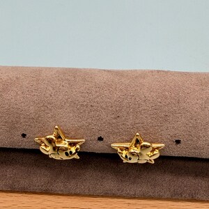 May include: A pair of gold-colored star-shaped earrings. Each earring features a small, detailed design within the star shape. The earrings are displayed on a brown surface with a light blue background.