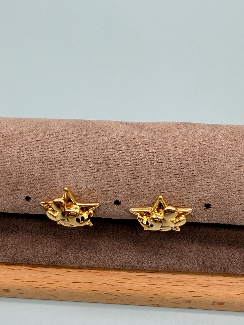 May include: A pair of gold-colored star-shaped earrings. Each earring features a small, detailed design within the star shape. The earrings are displayed on a brown surface with a light blue background.