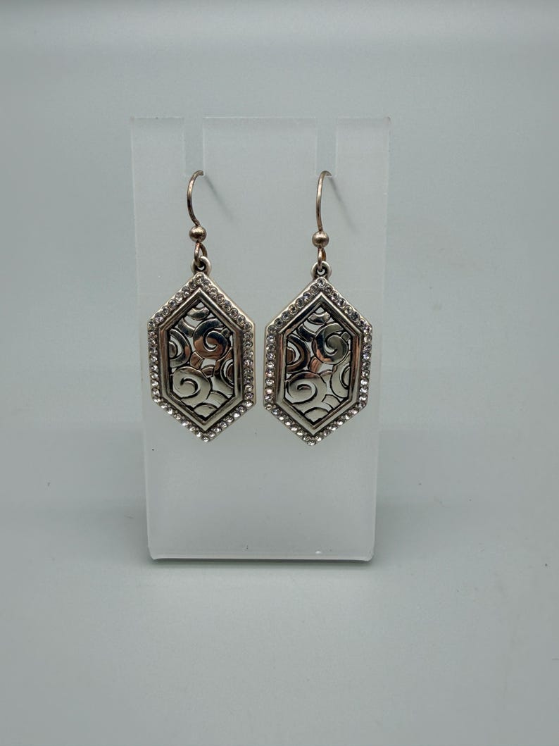 Puede incluir: Un par de pendientes plateados con forma hexagonal. Cada pendiente presenta un patr&oacute;n decorativo de remolinos en el centro, enmarcado por peque&ntilde;os cristales transparentes. Los pendientes cuelgan de un gancho simple.