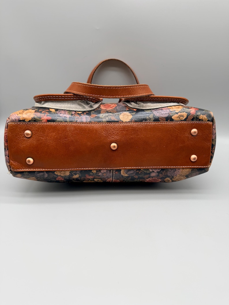 May include: A floral print handbag with brown leather accents. The bag features a top handle and a brown leather base with copper rivets. The floral pattern includes red, orange, and yellow flowers on a dark background.