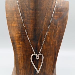 Pu&ograve; includere: Una collana color argento con un ciondolo a forma di cuore. Il ciondolo presenta un design a spirale ed &egrave; decorato con piccoli abbellimenti scintillanti. La collana &egrave; esposta su un busto di legno su sfondo bianco.