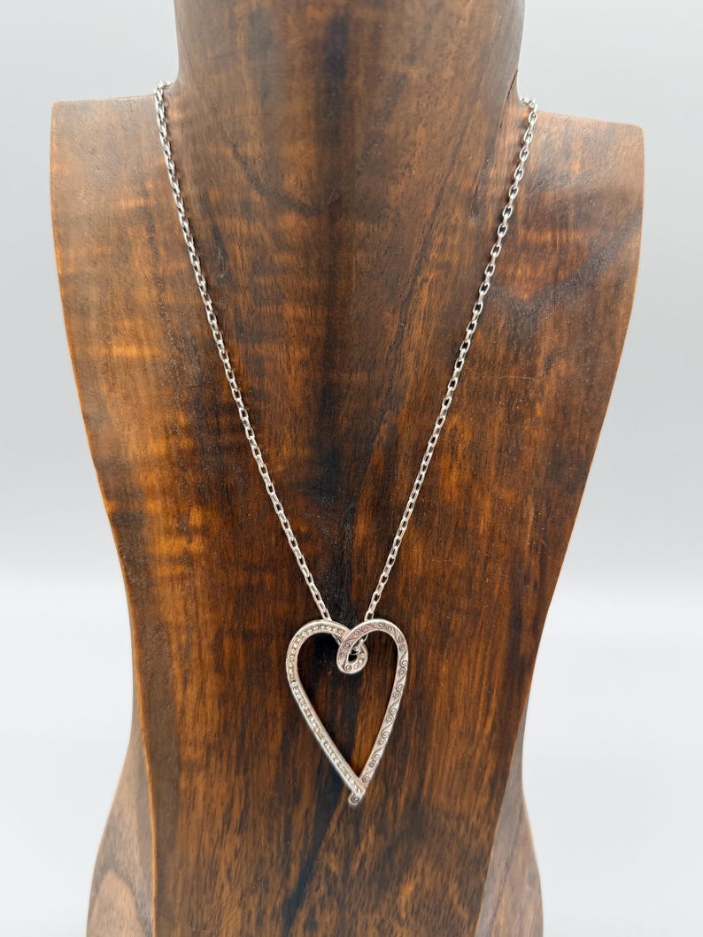 Pu&ograve; includere: Una collana color argento con un ciondolo a forma di cuore. Il ciondolo presenta un design a spirale ed &egrave; decorato con piccoli abbellimenti scintillanti. La collana &egrave; esposta su un busto di legno su sfondo bianco.