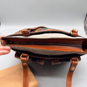 May include: Open view of a brown leather tote bag with a floral pattern. The bag has two handles, a zipper closure, and a light brown interior. The bag is approximately 12 inches wide and 10 inches tall.