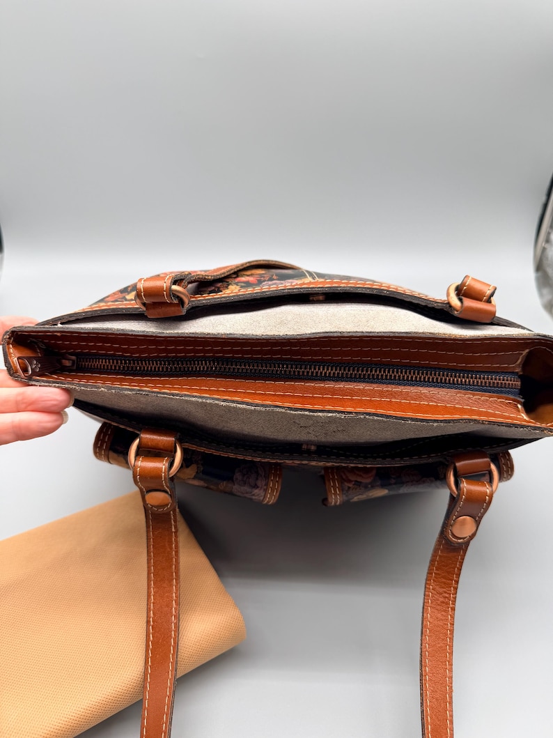 May include: Open view of a brown leather tote bag with a floral pattern. The bag has two handles, a zipper closure, and a light brown interior. The bag is approximately 12 inches wide and 10 inches tall.