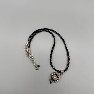 May include: A black beaded necklace with a silver clasp and a decorative circular pendant. The pendant features a central white stone surrounded by smaller black stones. The necklace is displayed on a light grey background.