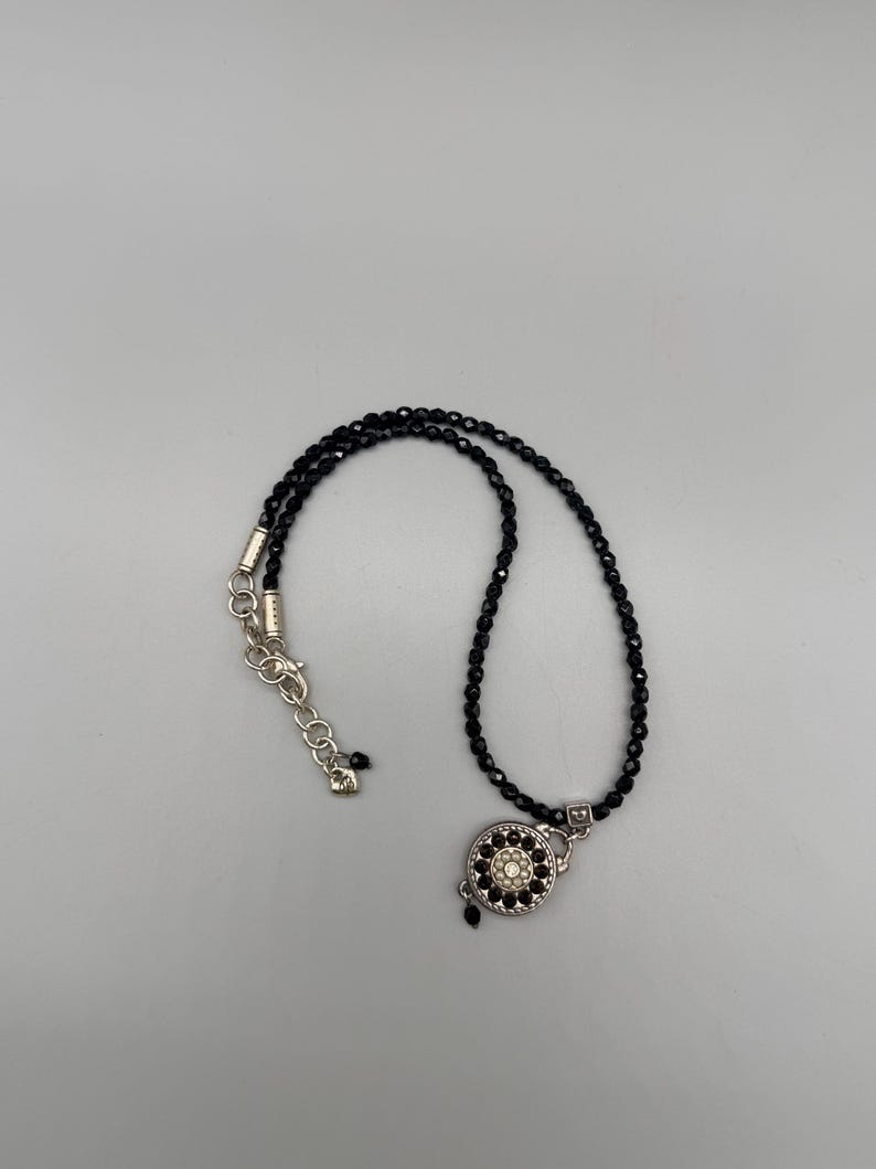 May include: A black beaded necklace with a silver clasp and a decorative circular pendant. The pendant features a central white stone surrounded by smaller black stones. The necklace is displayed on a light grey background.