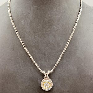 May include: A silver-toned rope chain necklace featuring a circular pendant. The pendant has a central clear round stone surrounded by a thin gold-toned ring and a textured silver-toned border with a twisted rope design and a decorative bail.