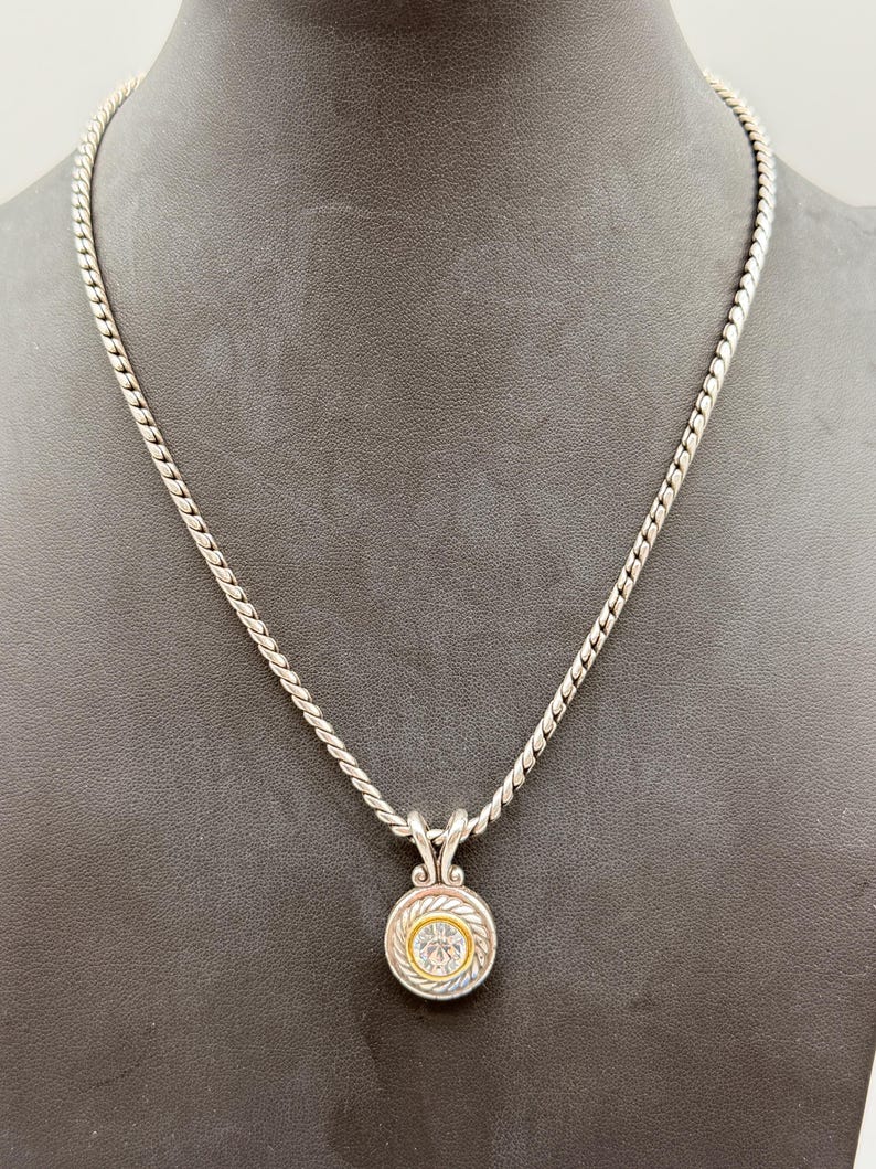 May include: A silver-toned rope chain necklace featuring a circular pendant. The pendant has a central clear round stone surrounded by a thin gold-toned ring and a textured silver-toned border with a twisted rope design and a decorative bail.