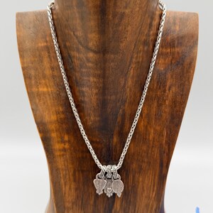 May include: A silver-toned wheat chain necklace featuring three small, textured, heart-shaped charms hanging from the center. The metal has an oxidized finish that highlights the intricate patterns on each charm, creating a detailed and cohesive look.