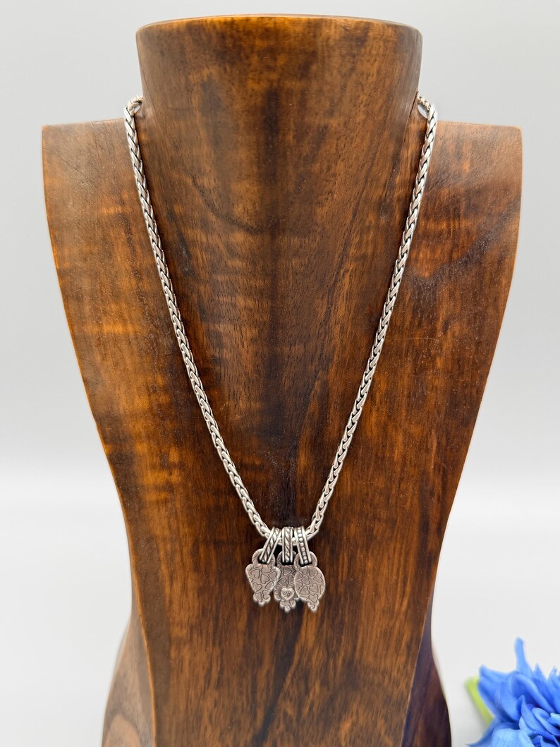 May include: A silver-toned wheat chain necklace featuring three small, textured, heart-shaped charms hanging from the center. The metal has an oxidized finish that highlights the intricate patterns on each charm, creating a detailed and cohesive look.