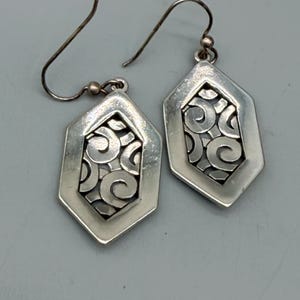 Puede incluir: Un par de pendientes plateados. Cada pendiente presenta una forma geom&eacute;trica hexagonal con un dise&ntilde;o calado decorativo de patrones arremolinados. Los pendientes tienen un cierre de gancho. El fondo es gris neutro.